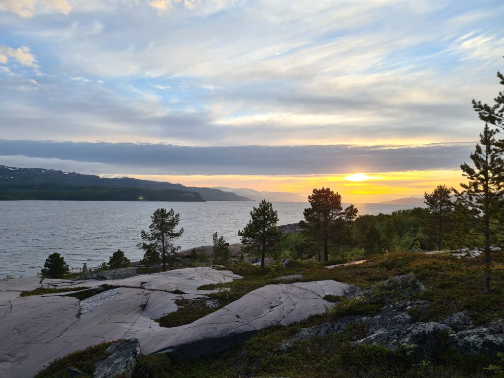 Fotot visar midnattssol över en fjord i fjällmiljö, i förgrunden finns kala hällar och småtallar.