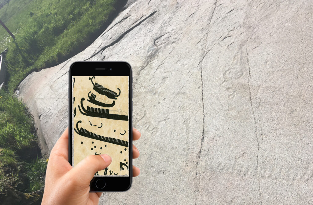Bilden visar en berghäll med ristningar. I förgrunden syns en hand med en smartphone.