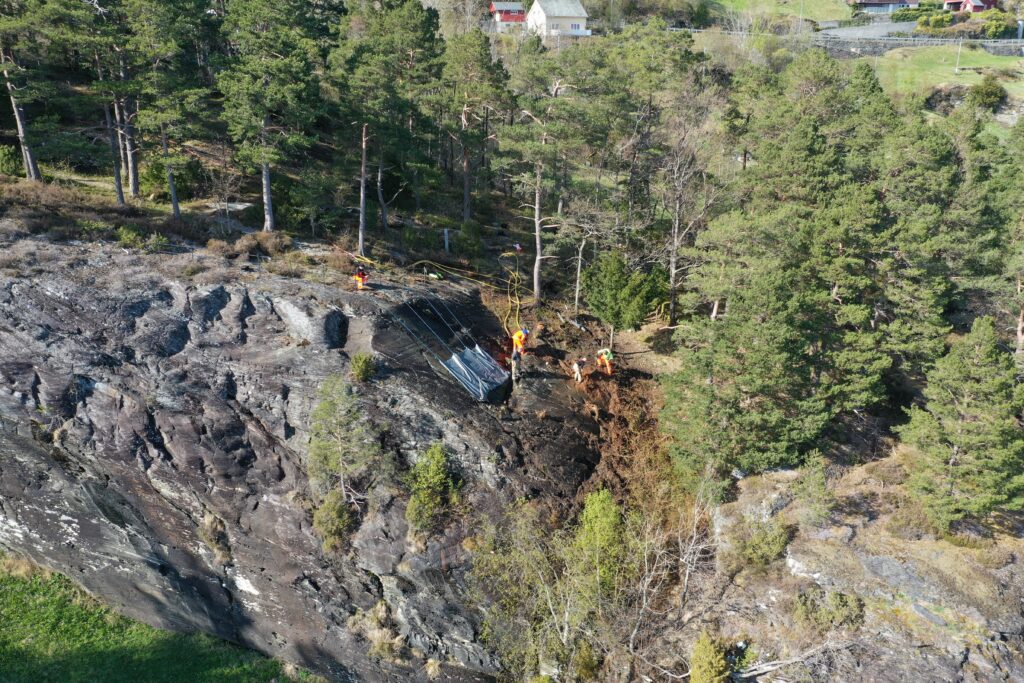 Fotot är taget uppifrån. Fyra personer arbetar på en berghäll. Hällen ligger högst upp på en större klippa. I bakgrunden syns tallskog.