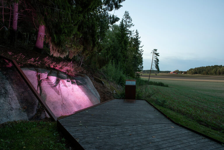 Fotot visar en häll med ristningar som belysts från sidan i skymningen. Ovanför ristningarna växer träd och till höger skymtas ett fält. Framför hällen finns en träramp.