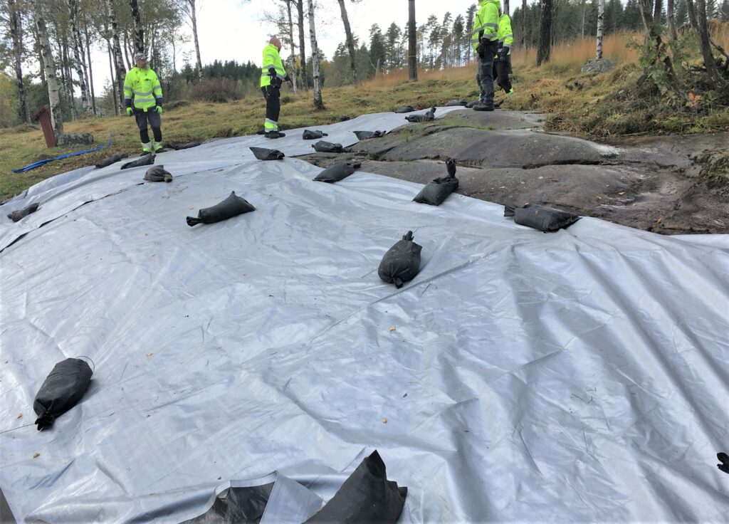 Fotot visar fyra personer vid en häll som är täckt med presenning, på den ligger sandsäckar.