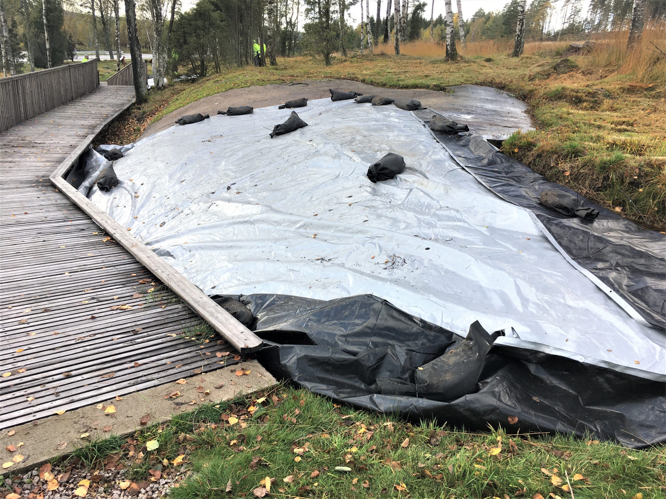 Fotot visar en täckt hällristningslokal. Hällen är täckt med presenning, på den ligger sandsäckar. Till vänster finns en träramp med räcke.