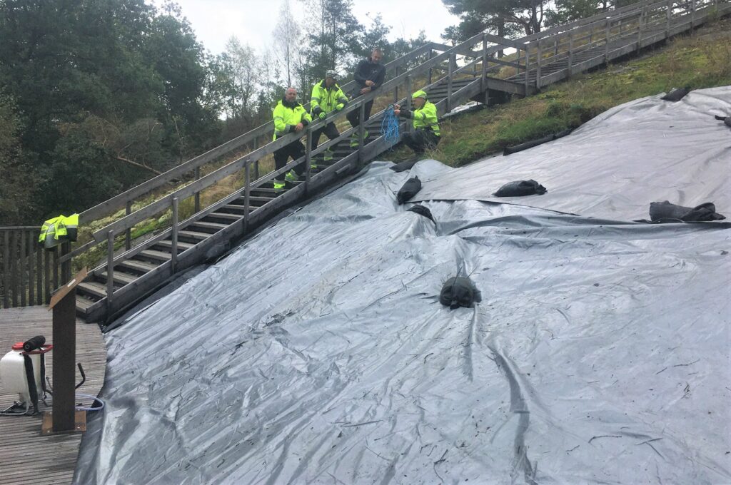 Fotot visar en täckt sluttande hällristningslokal. Hällen är täckt med presenning, på den ligger sandsäckar. Nedanför hällen syns ett trädäck med en skylt. I en trapp till vänster om hällen står fyra personer ur arbetslaget.