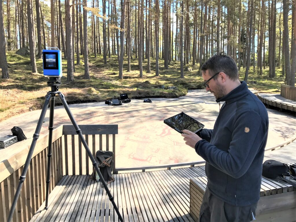 På fotot syns Kristofer Axelsson från MLT AB. Han håller i en pekplatta, till vänster står en landskapsskanner på ett stativ. I bakgrunden syns en berghäll med målade ristningar.