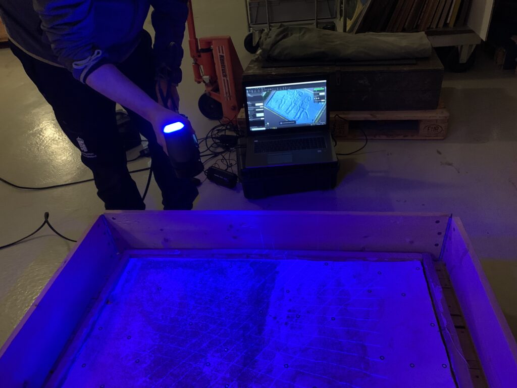 Fotot visar en gipsavgjutning i en pallkrage. Avgjutningen är upplyst av ett blått sken från den handhållna laserskannern.