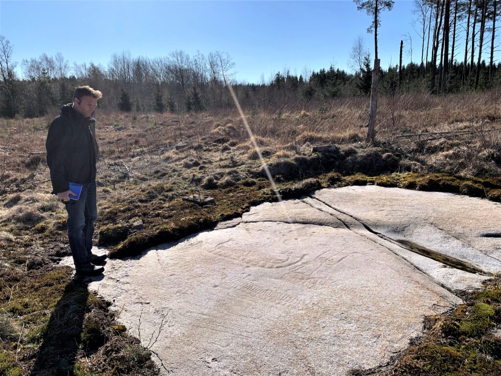 Fotot visar person som står vid en häll med ristningar.