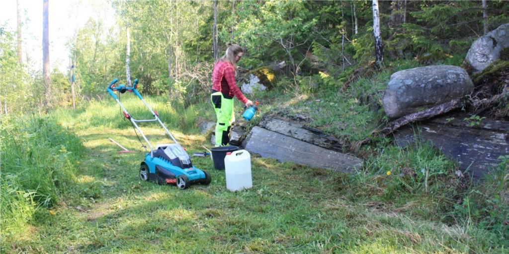 Fotot visar en person som sprejar etanol på en berghäll i ett skogsområde. Framför hällen står en dunk och en gräsklippare.