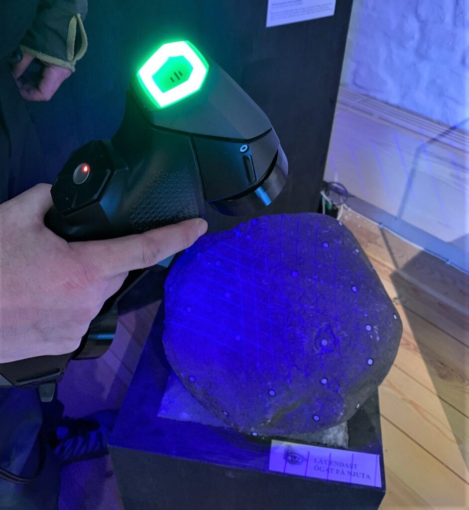 Fotot visar en sten som skannas med handhållen laserskanner, skannern lyser med ett blått sken på stenen.