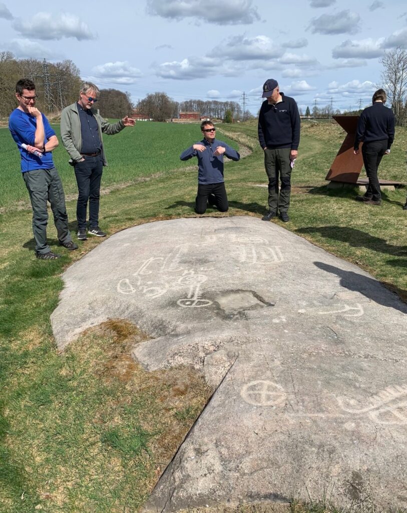 Foto på häll med ristningar i jordbruksmark, fem personer tittar på ristningarna..