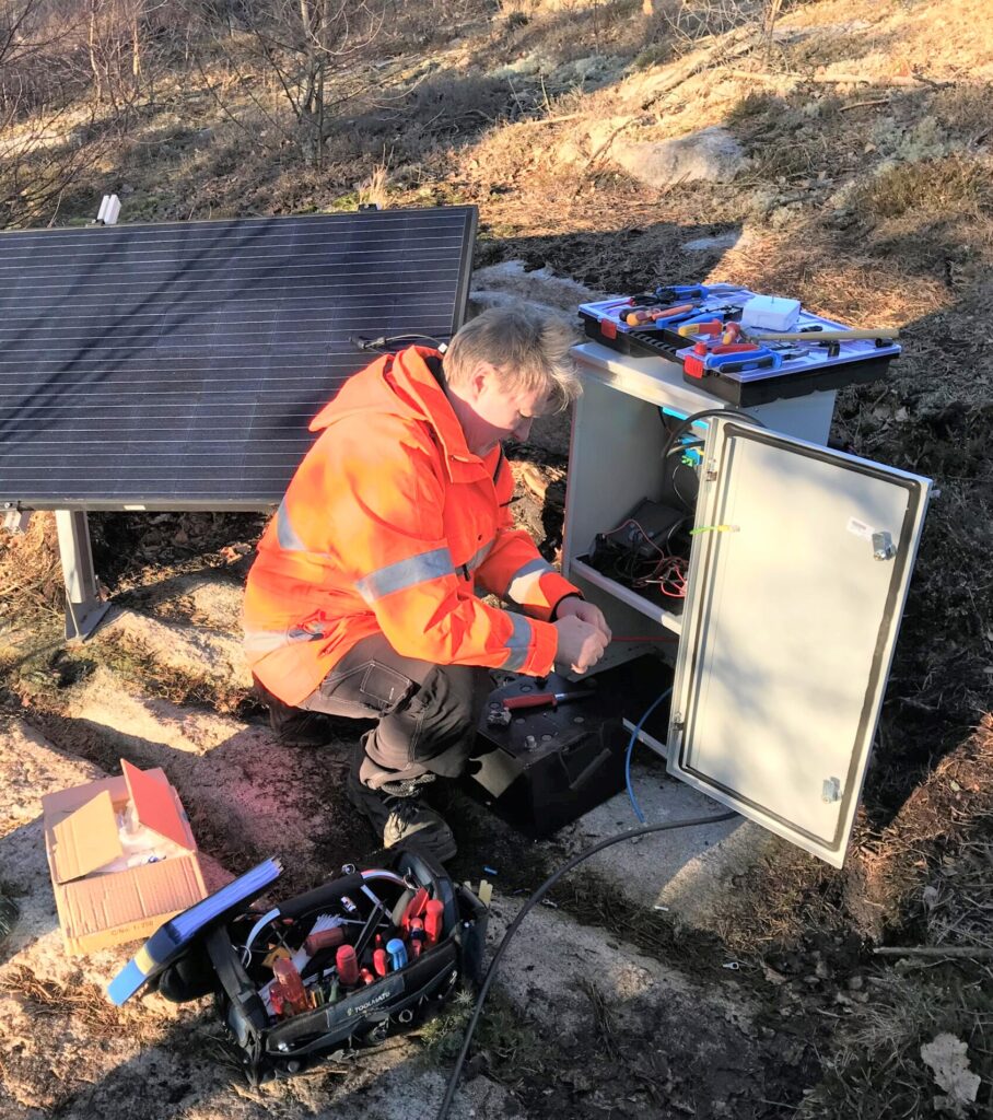 Fotot visar en person som sitter framför en elskåp och arbetar med installation av LED-belysning, i bakgrunden syns en solcellspanel.